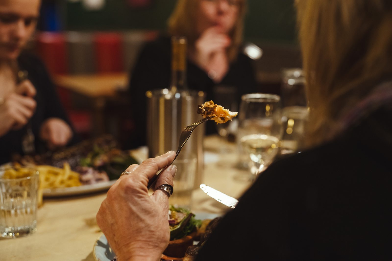 A table mid-meal, a forkful raised, friends gathered, glasses of wine